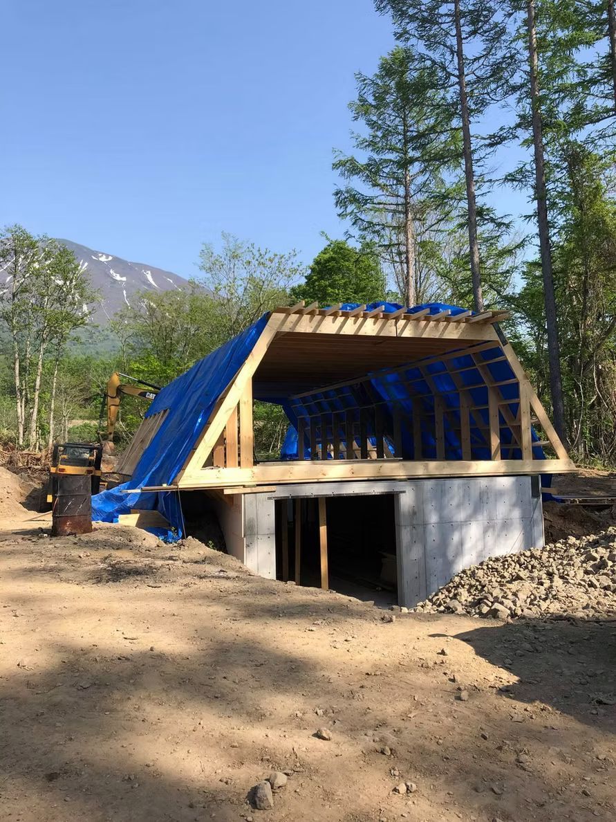 Eine im Bau befindliche Holzrahmenkonstruktion, die mit blauen Planen abgedeckt und auf einem Betonfundament in einem Waldgebiet errichtet wurde.