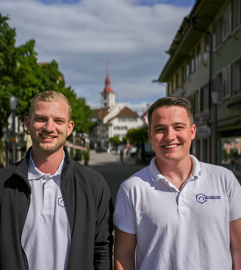 Zwei lächelnde Personen in weißen Poloshirts stehen auf einer Straße vor einem Stadtzentrum; im Hintergrund ist ein Kirchturm zu sehen.
