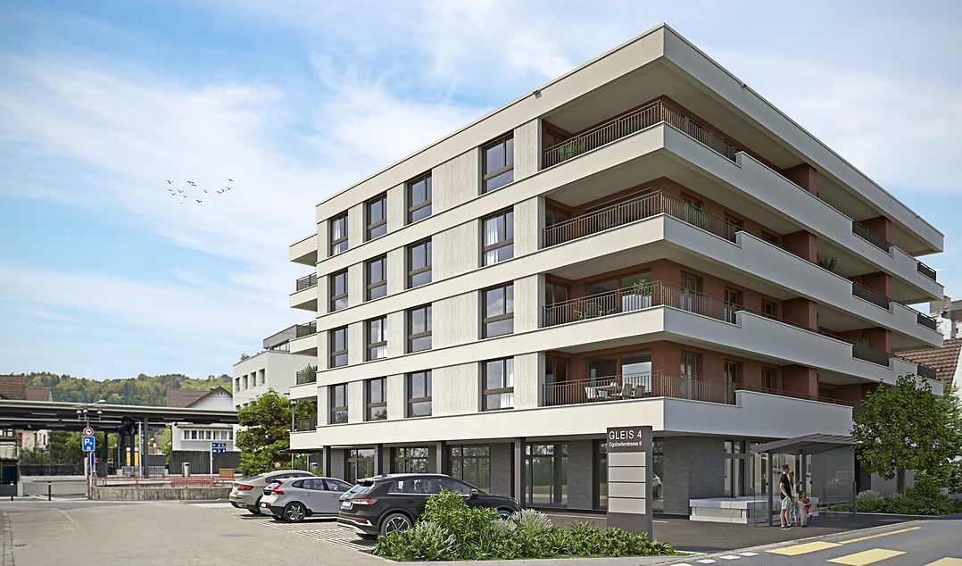 Ein modernes, mehrstöckiges Wohngebäude mit hellen Balkonen und Ladenlokalen im Erdgeschoss neben einem Parkplatz.