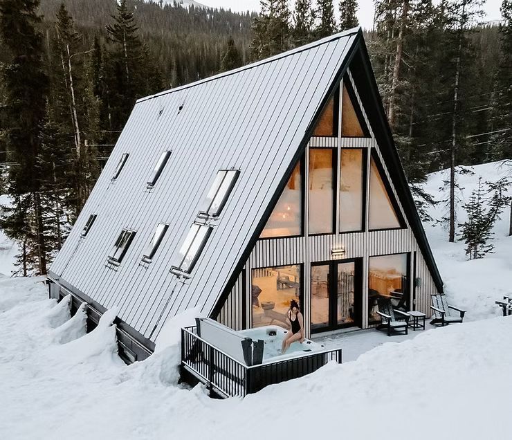 In einem verschneiten Wald liegt eine moderne A-förmige Hütte mit Metalldach, bodentiefen Fenstern und einem dampfenden Whirlpool.