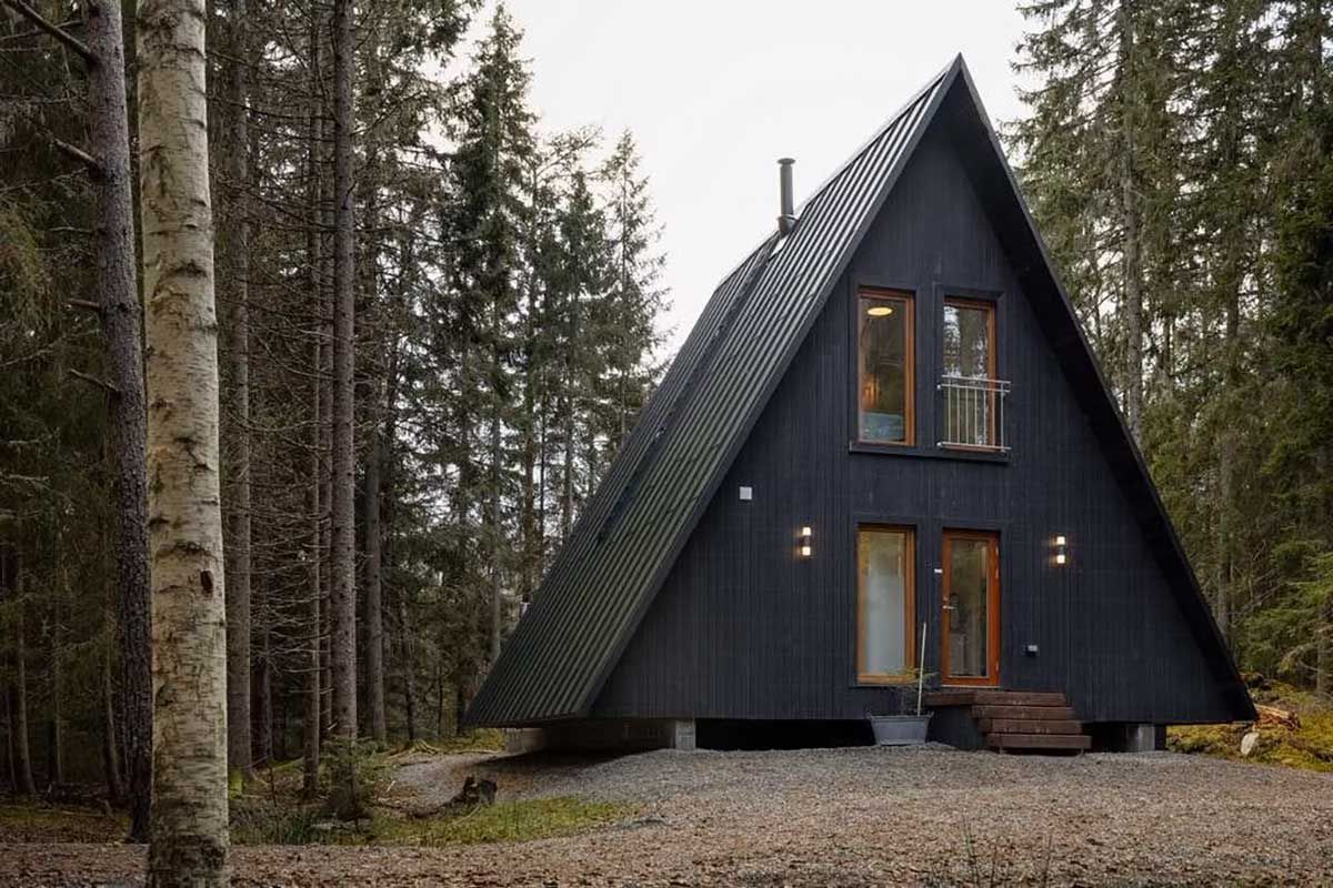 Eine moderne, schwarze A-förmige Hütte, eingebettet in einen Wald, mit Kiesboden und Holzstufen, die zum Eingang führen.