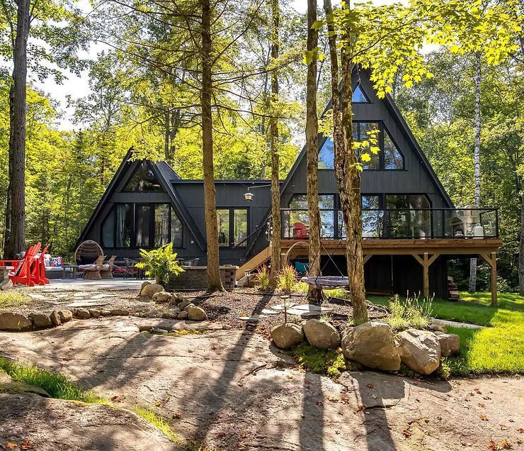 Ein dunkelgraues A-förmiges Haus, eingebettet in ein üppiges Waldgebiet, mit Steinterrasse, Holzdeck und hohen, sonnenbeschienenen Bäumen.