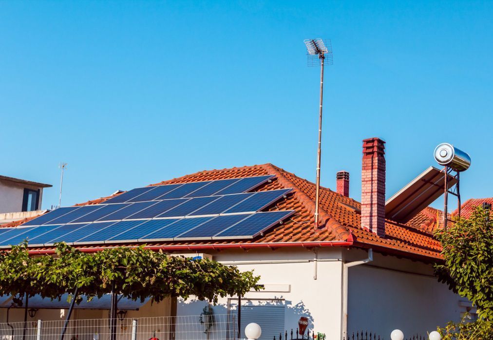 Paneles solares sobre un tejado de tejas rojas, con calentador de agua y antena. Cielo azul al fondo.
