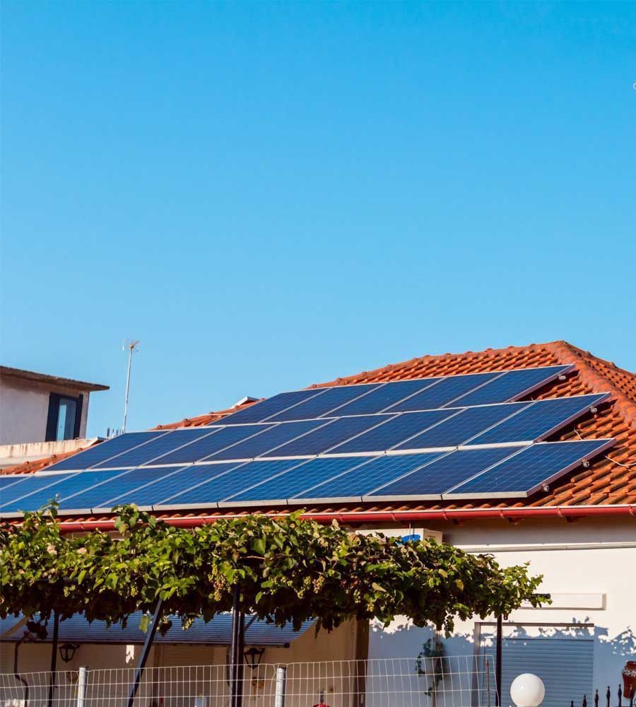 Paneles solares sobre un tejado de tejas rojas, bajo un cielo azul. Se ve una casa con árboles verdes en primer plano.