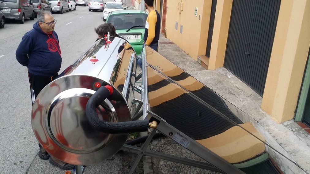 Personas examinando un gran calentador solar de agua en una calle de la ciudad. Tanque cromado, manguera negra y un panel para captar la luz solar.