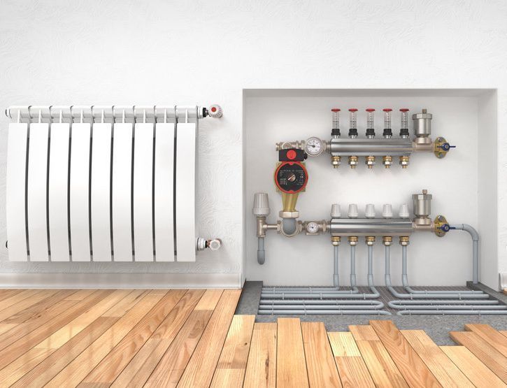 Instalación de radiadores blancos y sistema de calefacción por suelo radiante con tuberías, válvulas y suelo de madera sobre pared blanca.