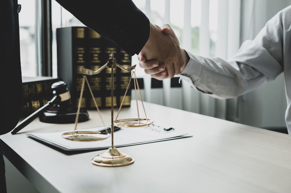 Dos personas se dan la mano sobre un escritorio con balanzas de justicia y libros de leyes.
