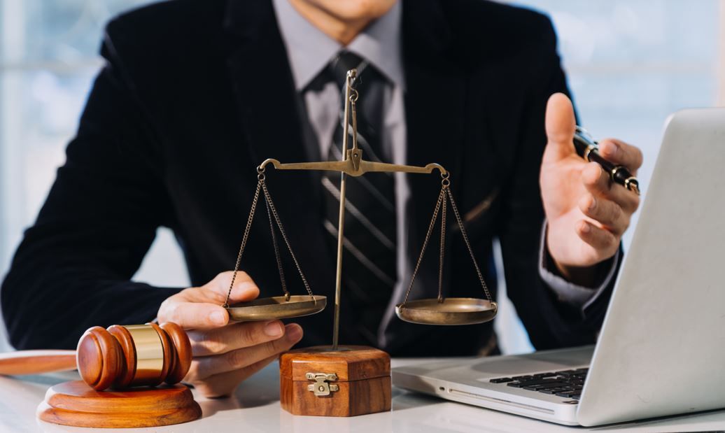 Persona de traje con mazo, balanza de la justicia y computadora portátil.