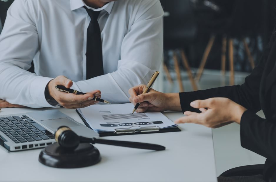 Dos personas vestidas de negocios revisando documentos, una firma, mazo sobre el escritorio.