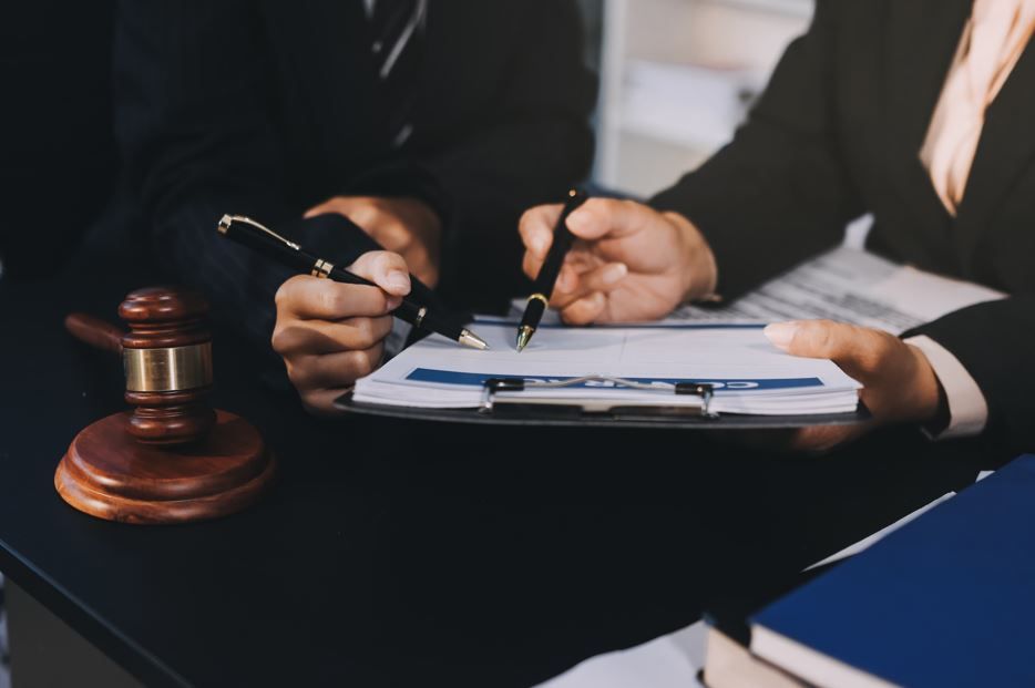 Dos personas trajeadas revisan documentos; una sostiene un bolígrafo cerca de un portapapeles. Un mazo reposa sobre la mesa.