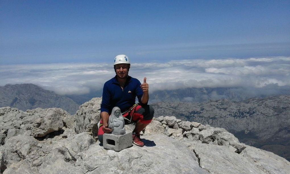 Un hombre está sentado en la cima de una montaña haciendo un gesto de aprobación con el pulgar.