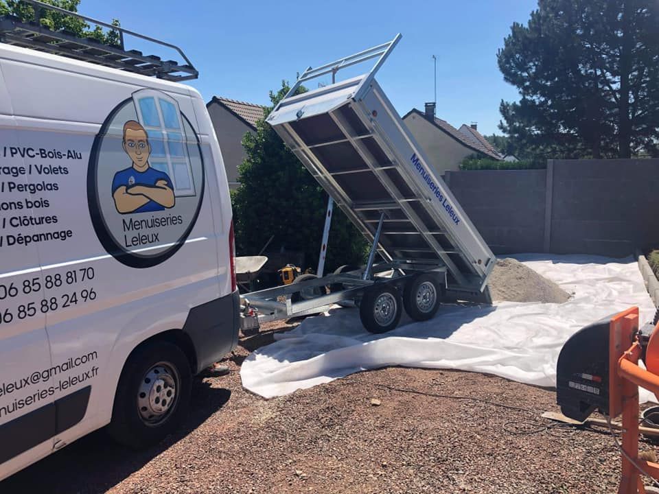 Une camionnette blanche avec remorque décharge du gravier sur une bâche dans une allée. La benne est surélevée et un panneau solaire est installé dessus.