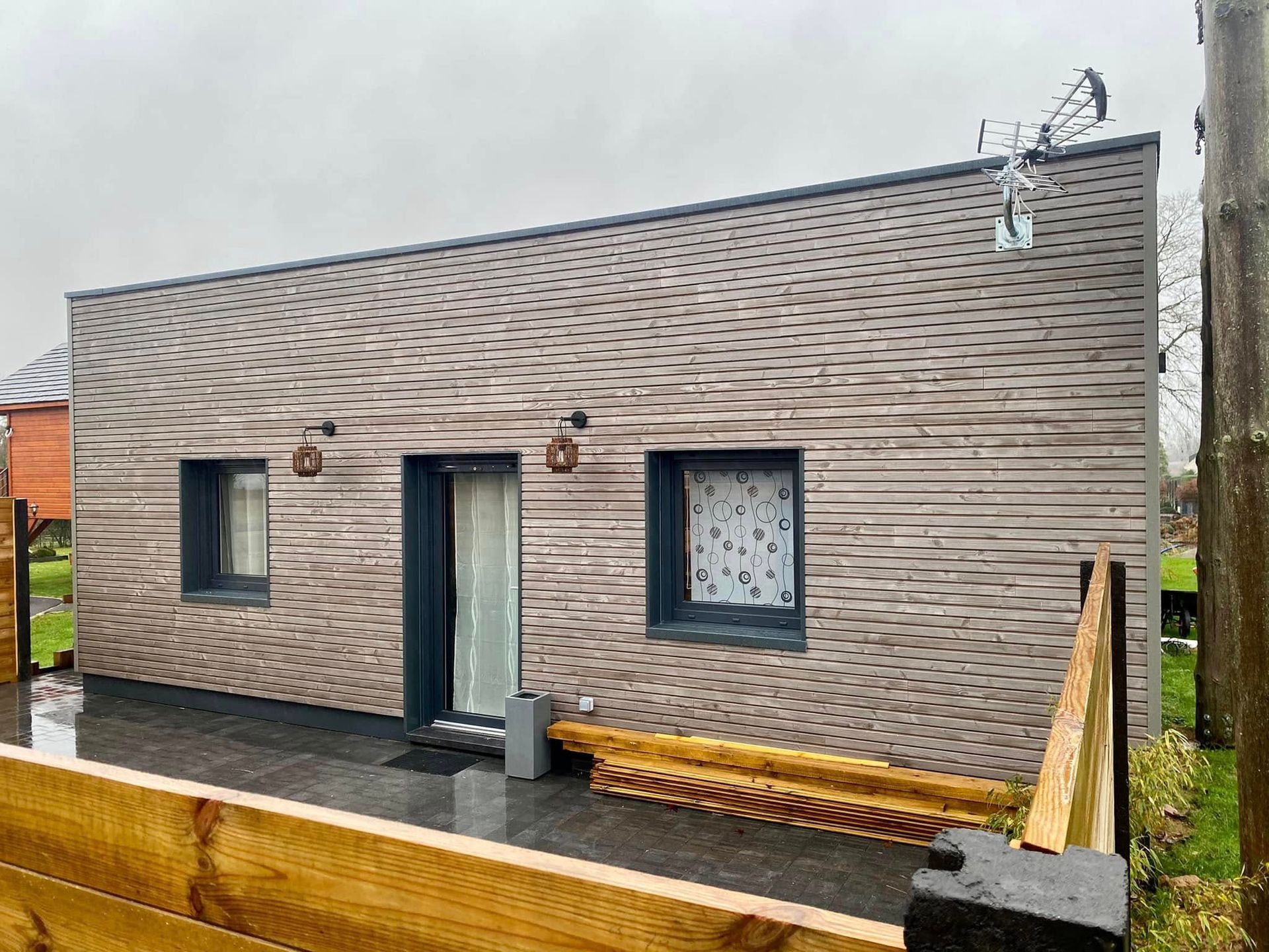 Petite maison moderne avec un bardage gris à motifs, deux fenêtres et une porte vitrée donnant sur une terrasse humide.