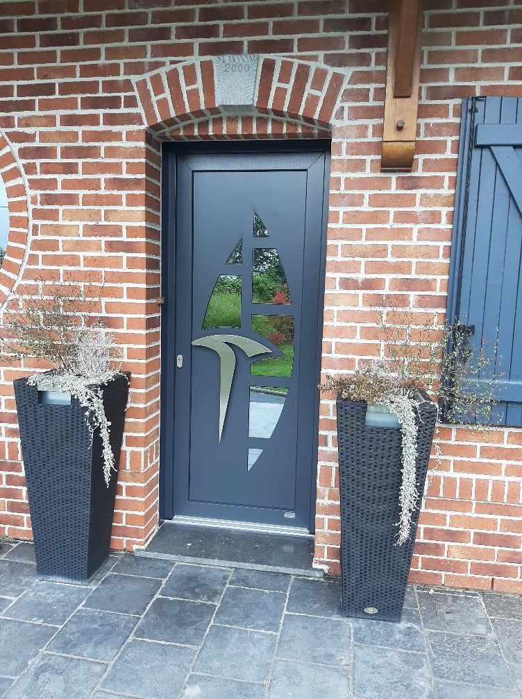 Entrée en briques avec porte bleu foncé, encadrement en briques cintrées et deux grands bacs à fleurs noirs sur une terrasse en pierre.