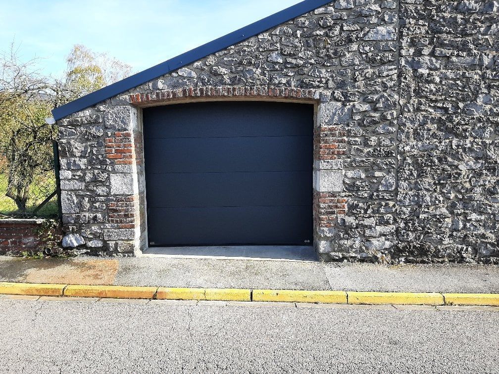 Bâtiment en pierre avec une porte de garage sombre et fermée, situé à côté d'un trottoir et d'une route.