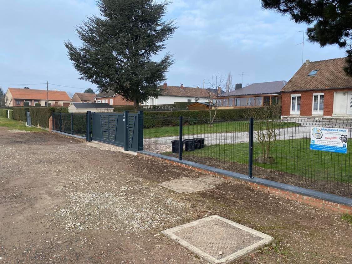 Allée et jardin résidentiels clôturés avec chemin de gravier, maisons en briques et un grand arbre sous un ciel nuageux.