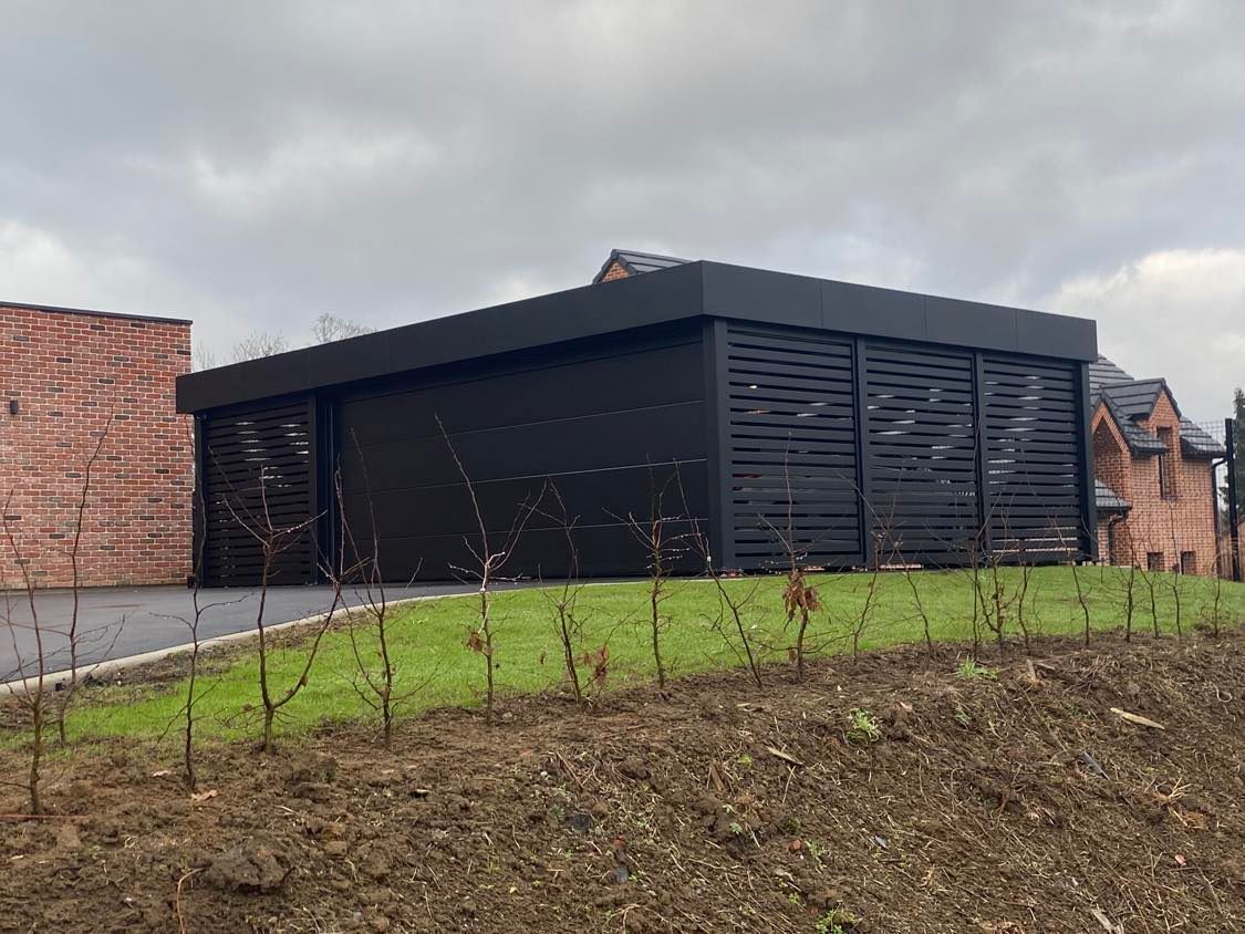 Bâtiment noir moderne sur une pente herbeuse sous un ciel nuageux, avec des maisons en briques en arrière-plan.