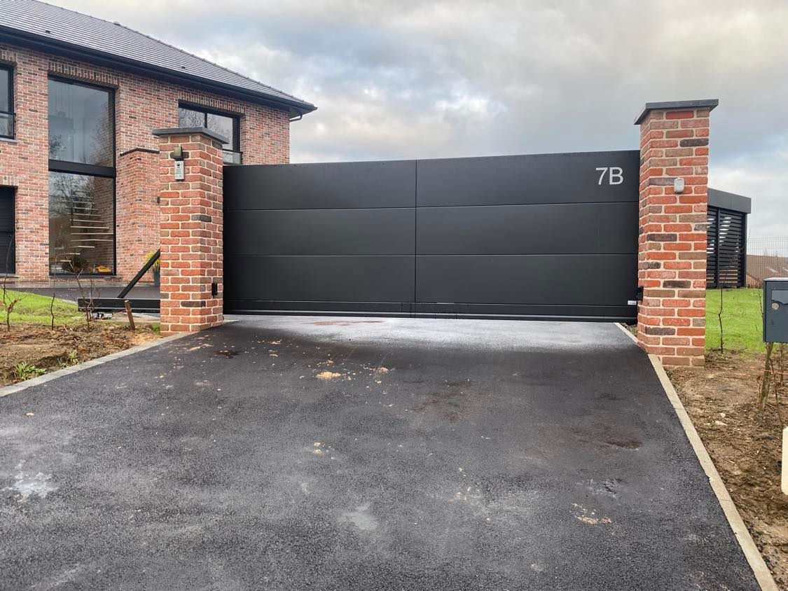 Portail double moderne noir entre des piliers en briques, à côté d'une maison, avec une allée pavée.