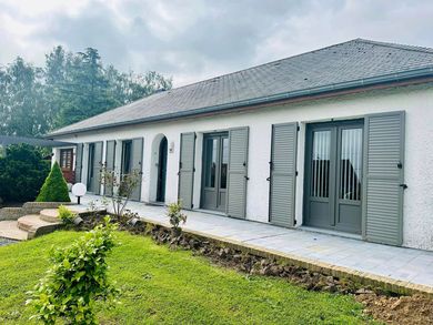 Maison moderne de plain-pied avec toit de tuiles grises, murs blancs et volets gris, donnant sur une pelouse verte.
