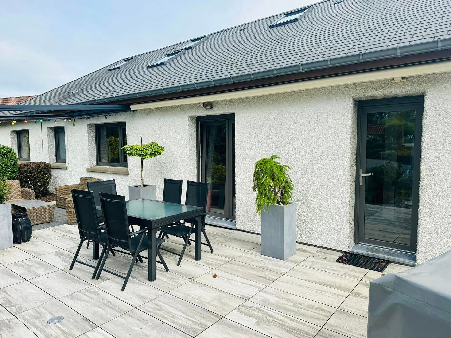 Terrasse avec table à manger noire, chaises, plantes en pot et façade d'une maison en pierre claire avec toit de tuiles foncées.