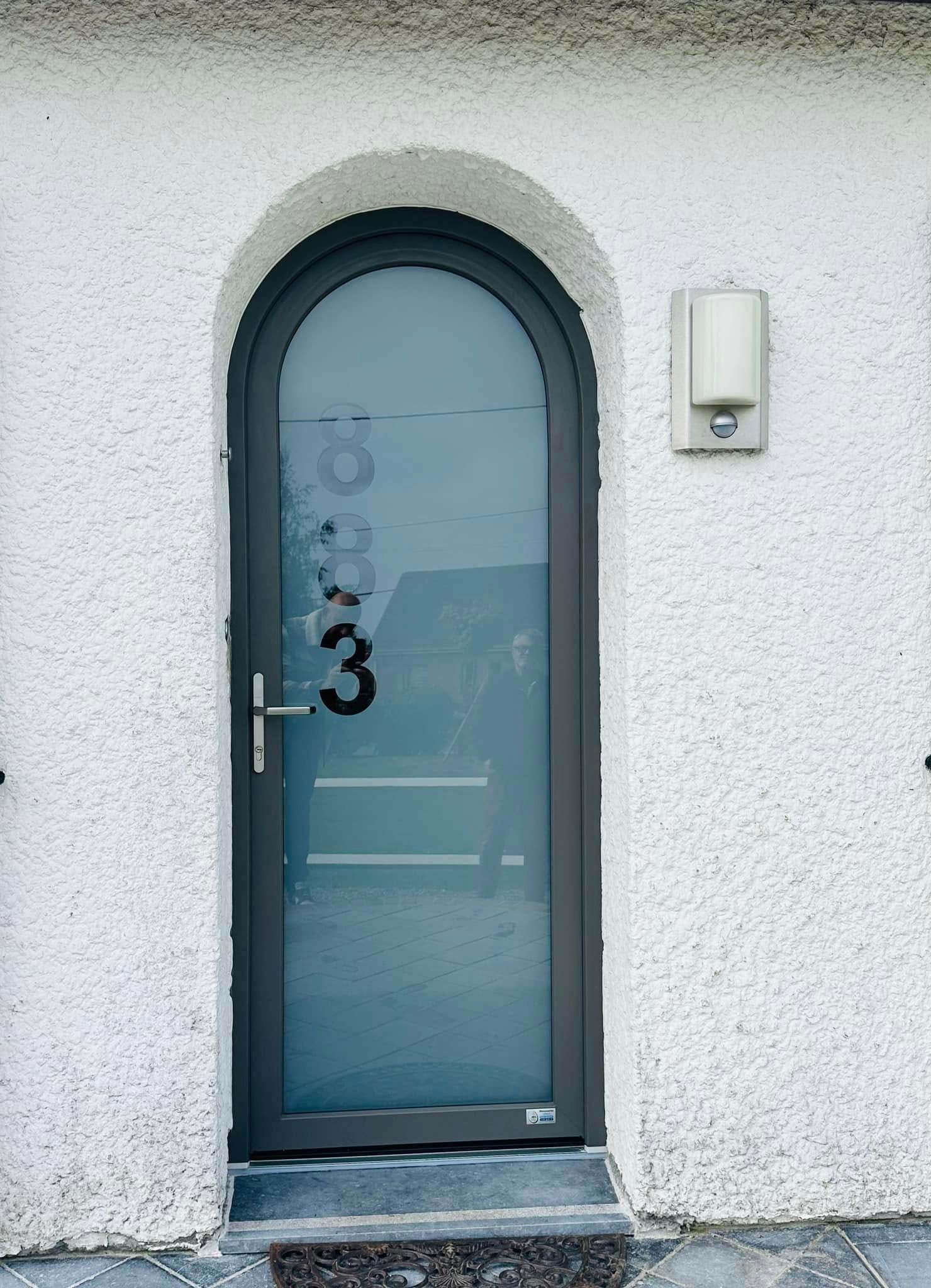 Porte d'entrée cintrée bleu-gris dans un mur en stuc blanc, avec une petite applique murale à côté.