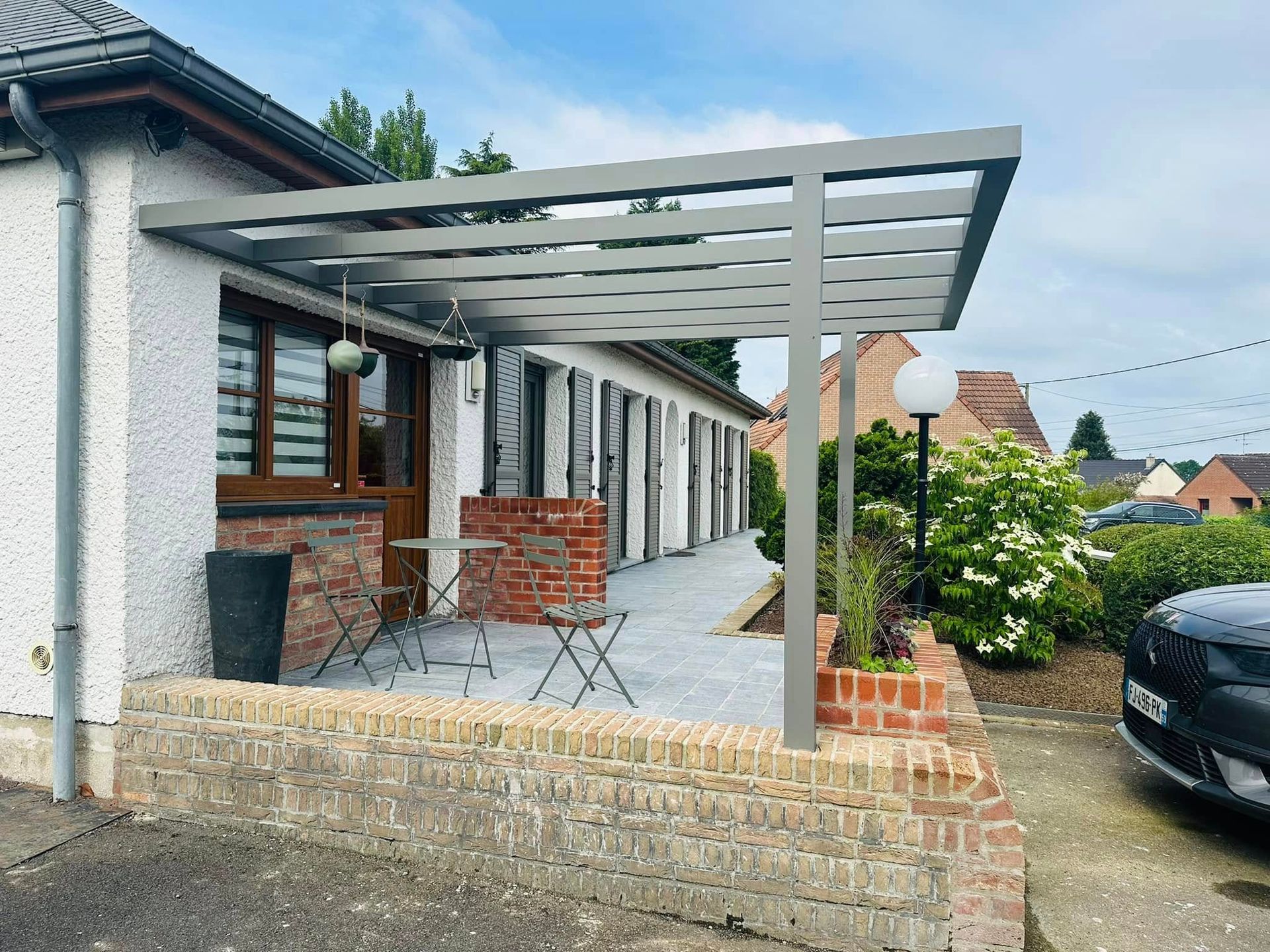 Patio avant avec pergola grise, piliers en briques et allée pavée longeant une maison.