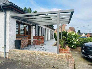 Patio d'une maison moderne avec pergola grise, jardinières en briques et voiture garée contre un mur en pierre.