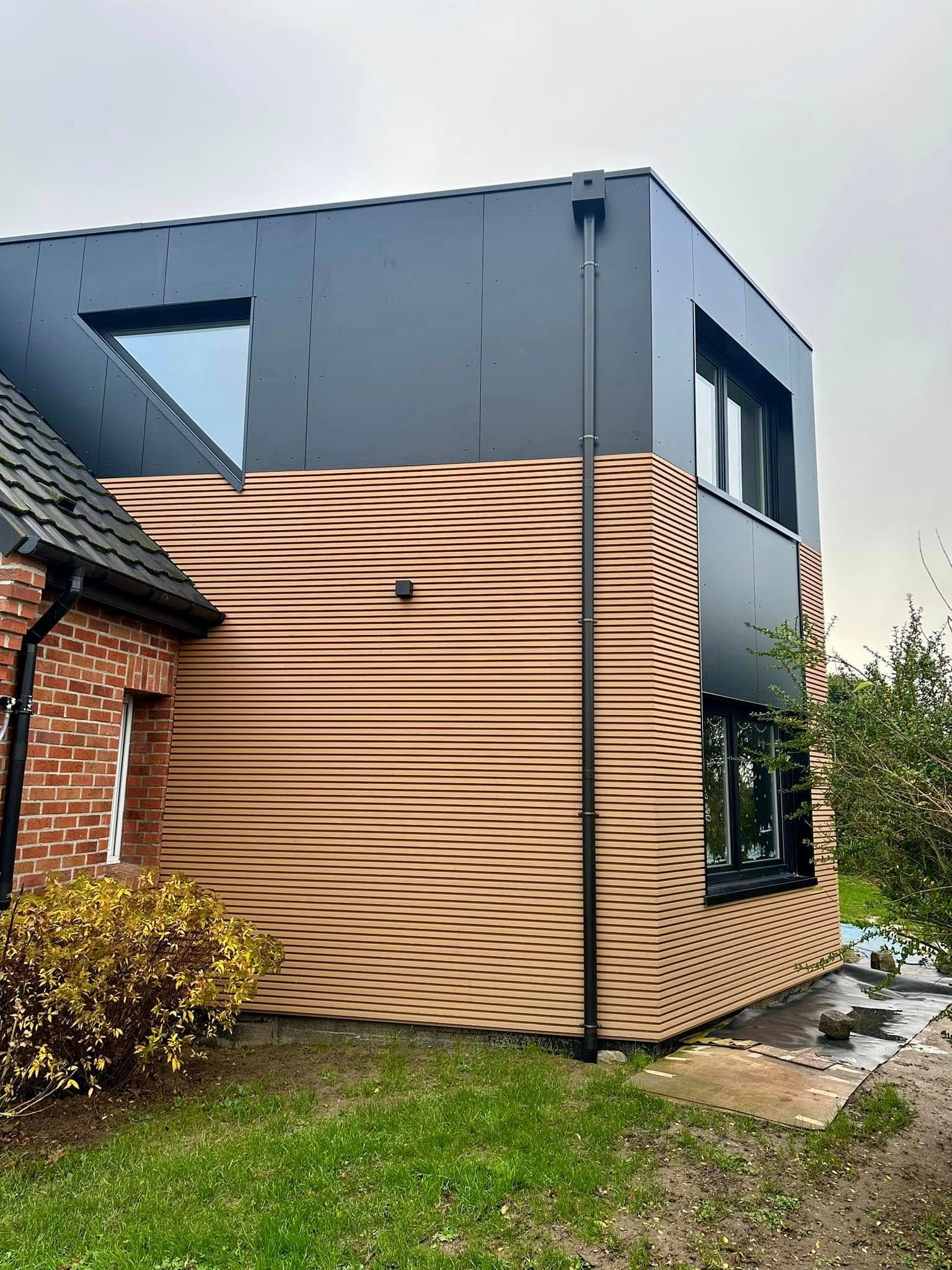 Extérieur d'une maison moderne à deux étages avec un revêtement foncé, un bardage en bois et un mur de briques donnant sur une cour gazonnée.