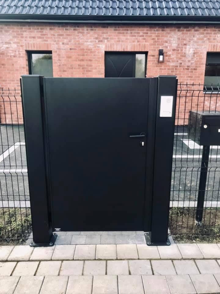 Portail et porte en métal noir devant un bâtiment en briques, avec un panneau à clavier sur la droite.
