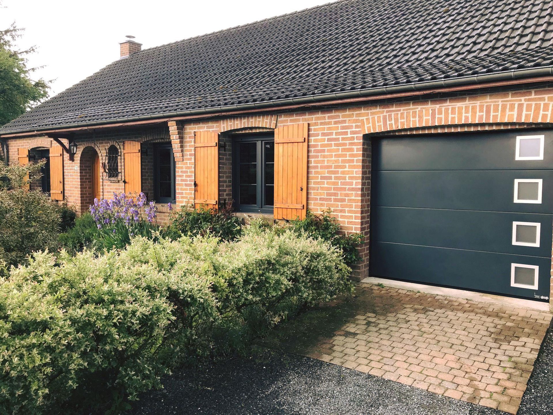 Maison en briques avec porte de garage noire, haie et allée pavée.