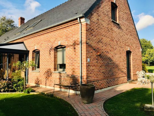 Maison en briques au toit sombre, petite terrasse et pelouse verte dans un jardin ensoleillé.