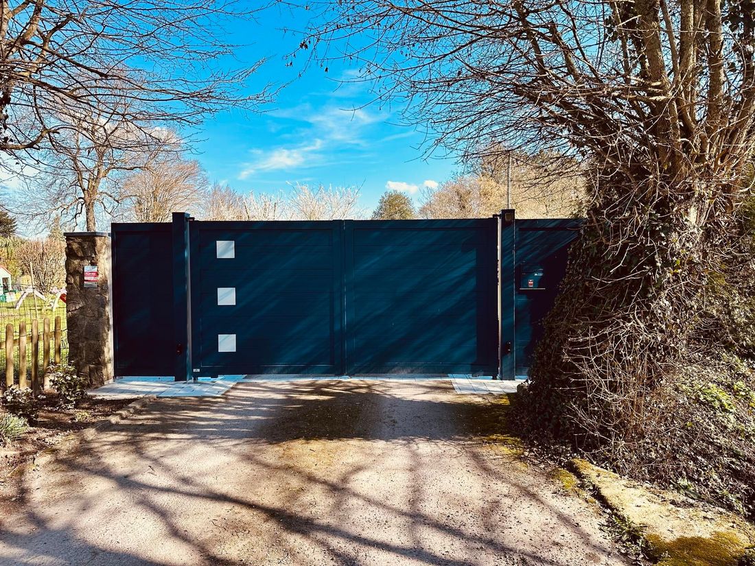 Un portail noir barrant une allée, encadré d'arbres et de haies par une journée ensoleillée.