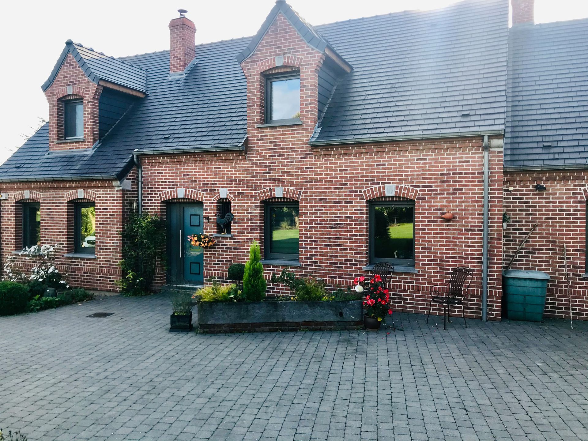 Maisonnette en briques avec une porte turquoise, des lucarnes, des jardinières et une allée pavée.
