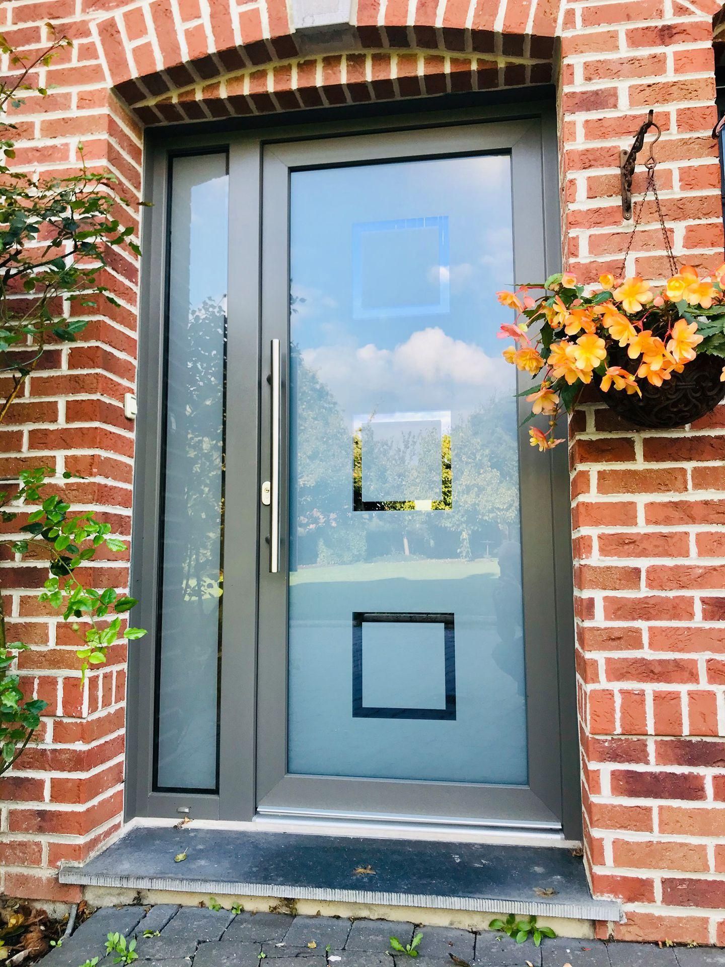 Entrée principale en briques avec porte grise, panneaux de verre dépoli et fleurs orange dans une jardinière murale.