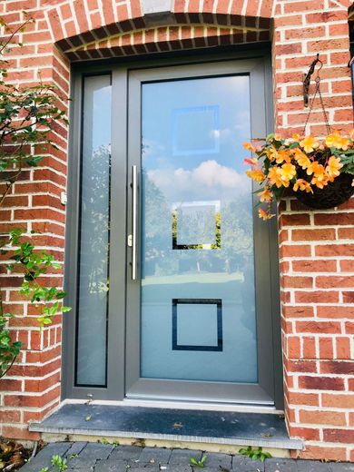Entrée principale en briques avec porte grise, panneaux de verre dépoli et fleurs orange dans une jardinière murale.