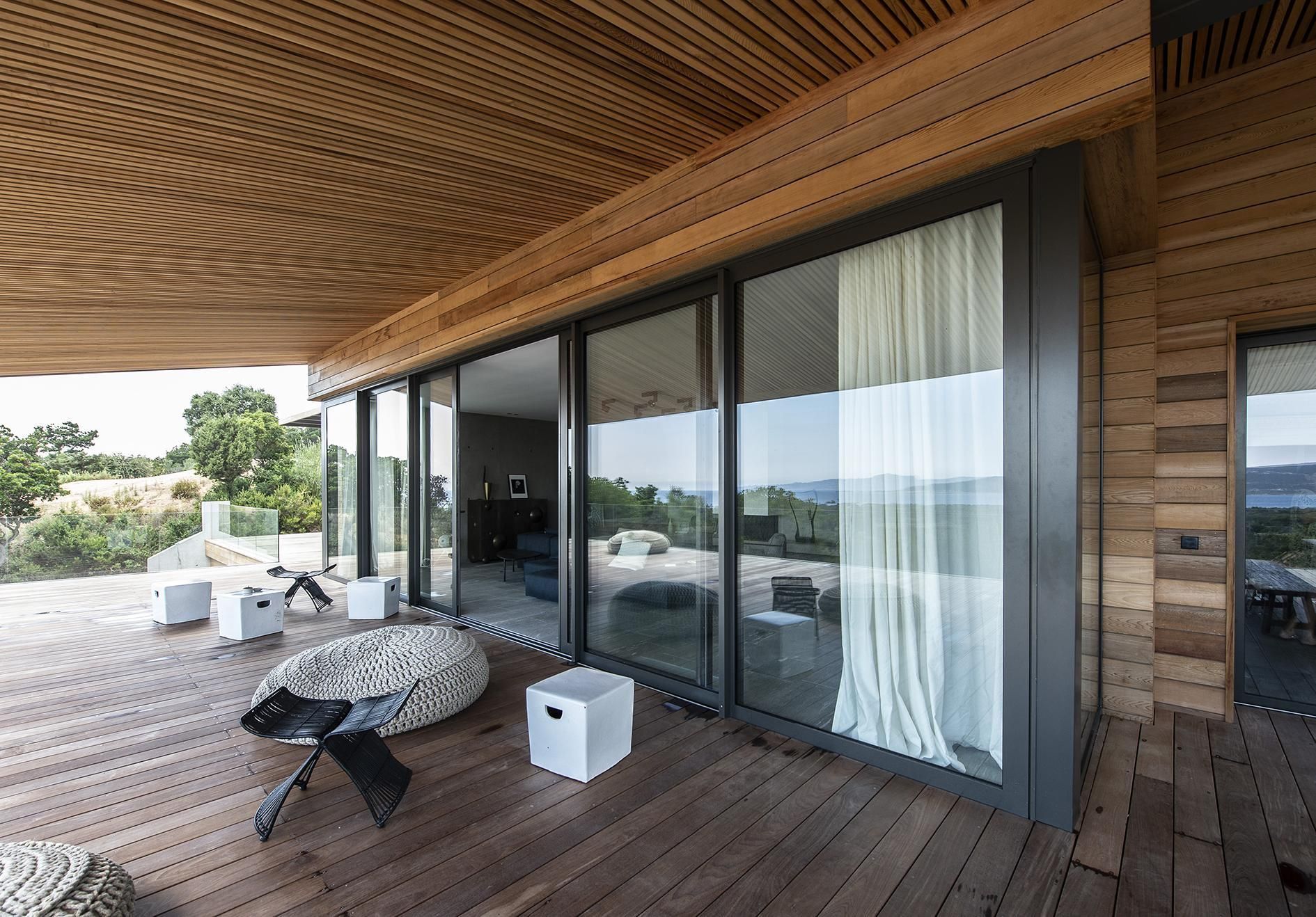 Terrasse moderne en bois avec plafond incurvé, portes vitrées, table d'extérieur, chaises et rideaux blancs.
