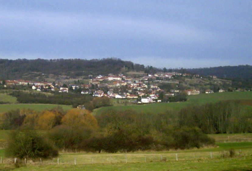 Village lointain sur une colline verdoyante sous un ciel bleu nuageux, avec des champs et des arbres au premier plan.