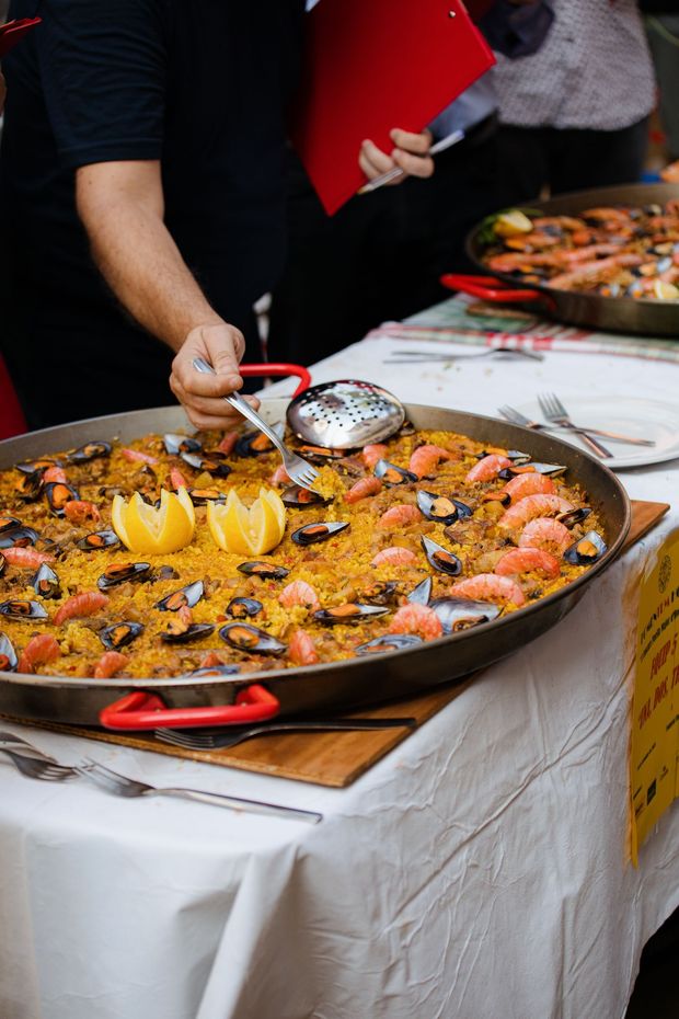 Une grande paella servie en plein air, avec des crevettes, des quartiers de citron et une louche posée sur une table.