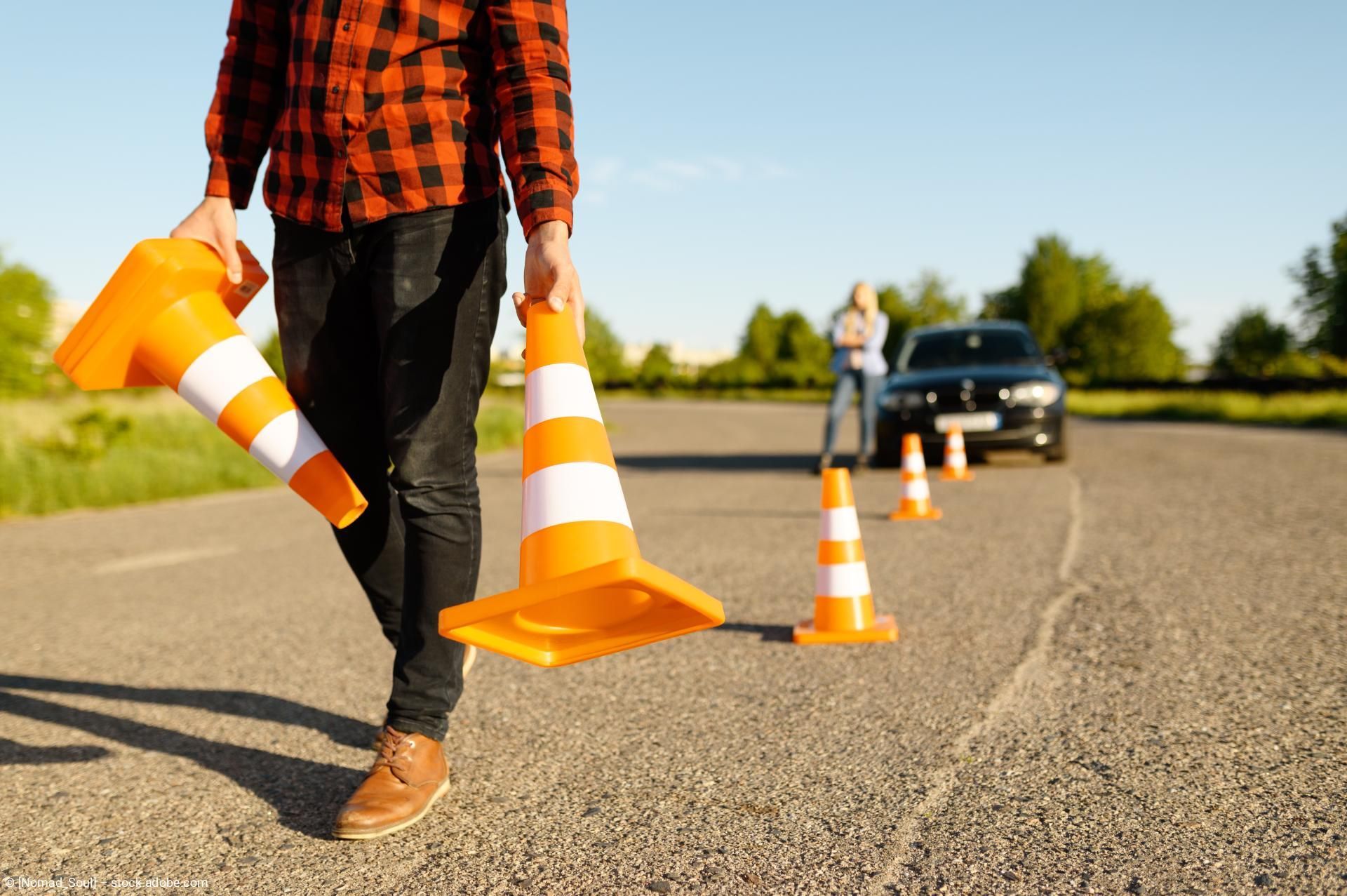 Ein Arbeiter in einem orange-karierten Hemd trägt Verkehrskegel auf einer Straße; im Hintergrund ist ein Auto zu sehen.