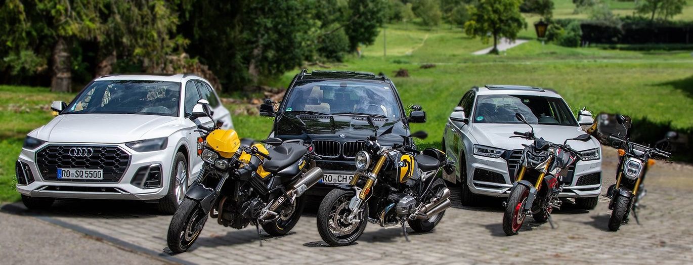 Ein weißer SUV und eine schwarze Limousine parken hinter drei Motorrädern am Straßenrand; im Hintergrund sind Bäume zu sehen.