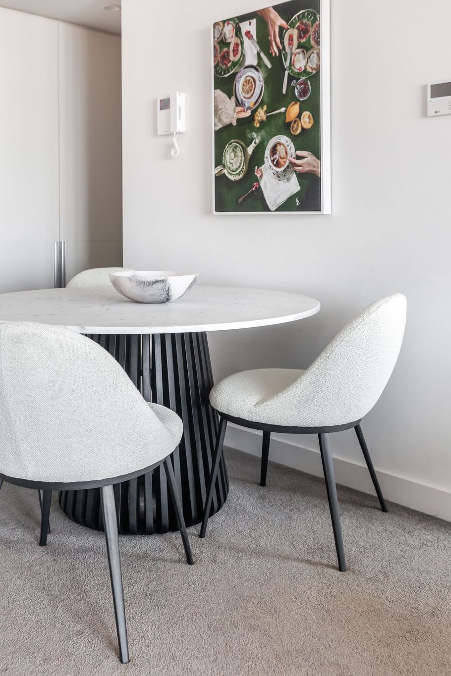 Small round dining table with white woven chairs in a bright minimalist room, under a floral wall art piece.