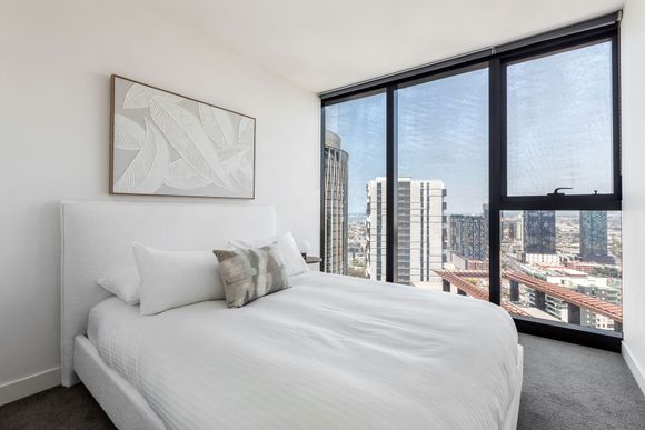 Bright bedroom with a bed and large windows overlooking a city skyline.