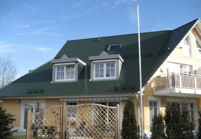 Gelbes Haus mit grünem Dach, Dachgauben und einem hohen weißen Mast unter blauem Himmel.
