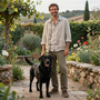 Un hombre de pie en un sendero del jardín junto a un perro negro, con flores y muros de piedra al fondo