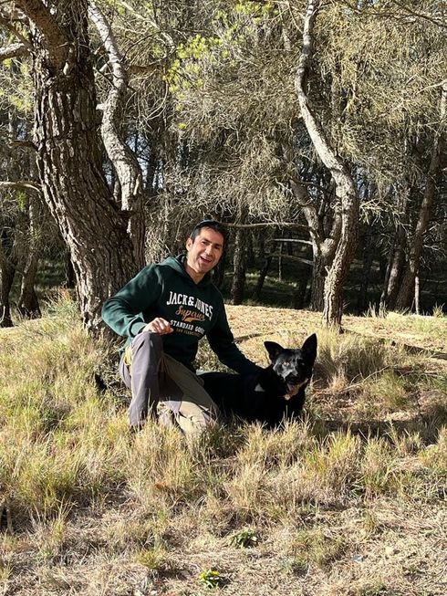 Persona sentada en la hierba junto a un perro negro en una zona boscosa y soleada