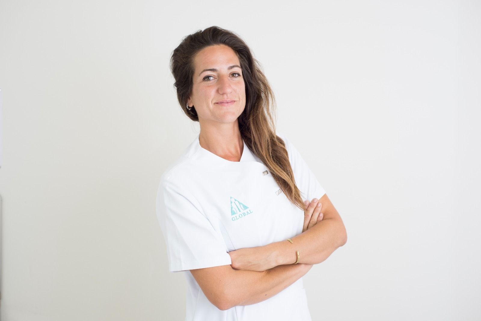 Mujer con los brazos cruzados, con una camisa blanca con un logo, frente a una pared blanca. Sonriendo.