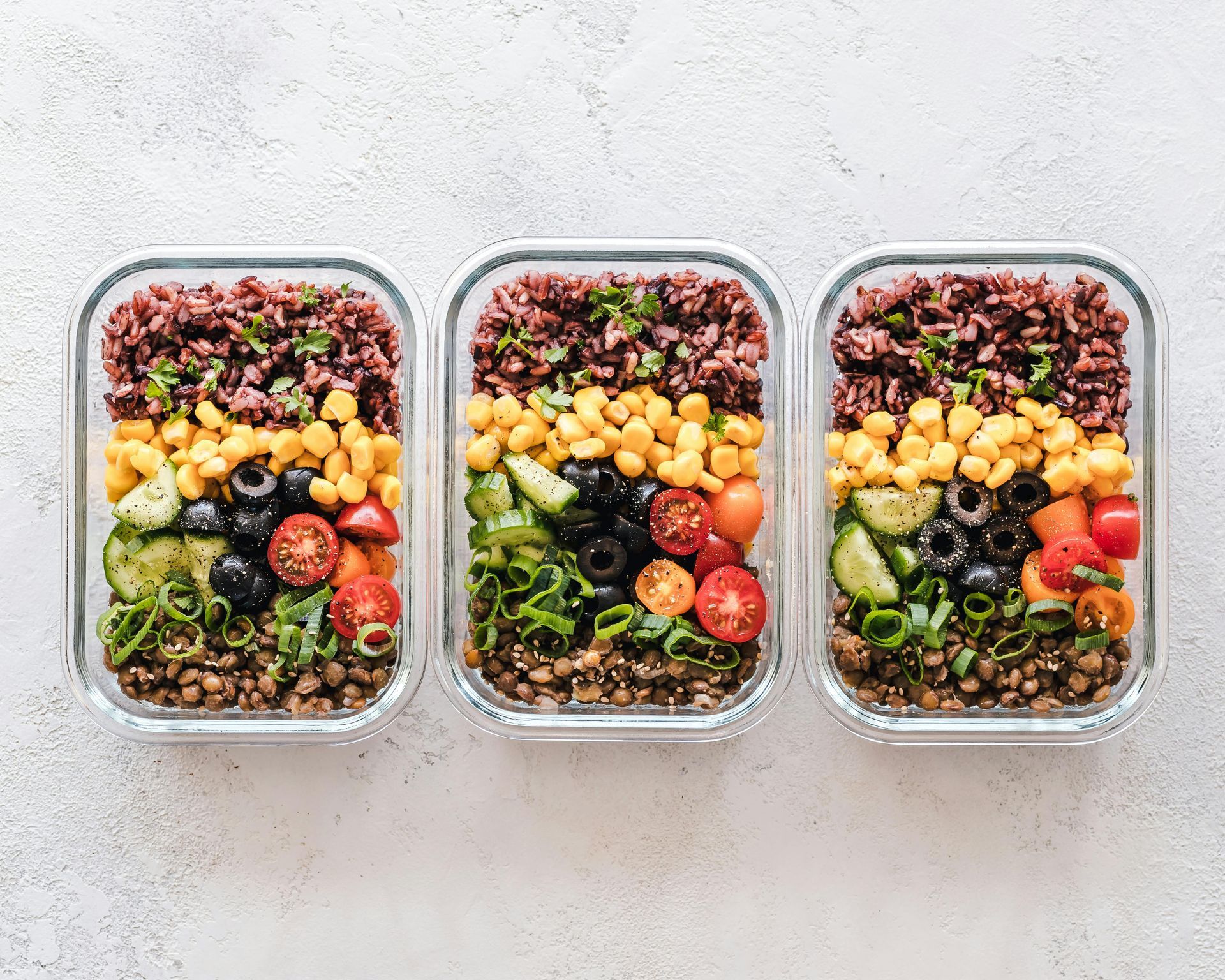 Tres recipientes para preparar comidas llenos de capas de granos, maíz, aceitunas, tomates y verduras.