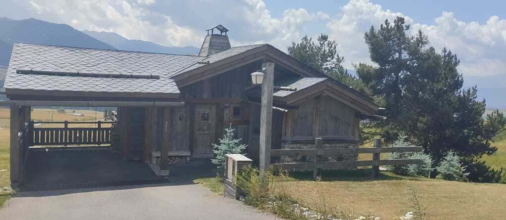 Cabaña rústica de madera con chimenea de piedra y cochera, situada en un paisaje rural soleado.