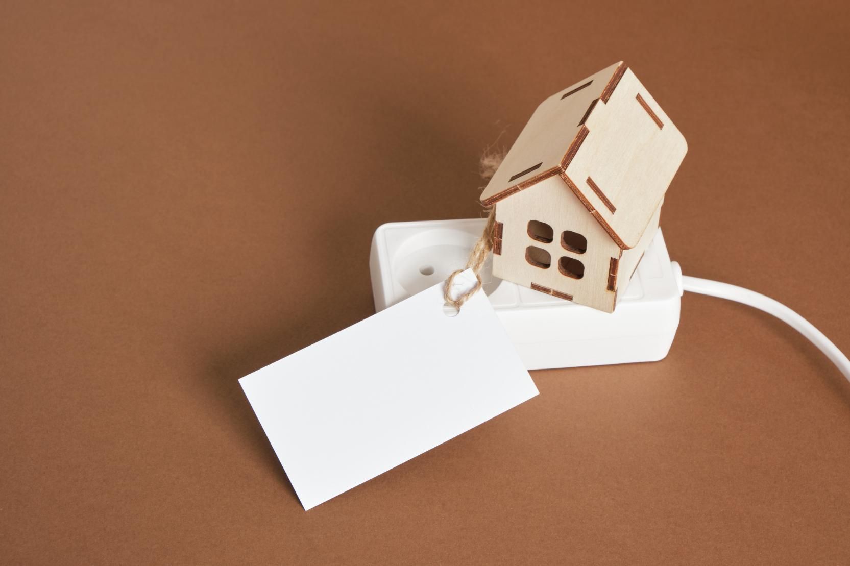 Maqueta de casa de madera conectada a una regleta, con una etiqueta blanca vacía. Sobre un fondo marrón.