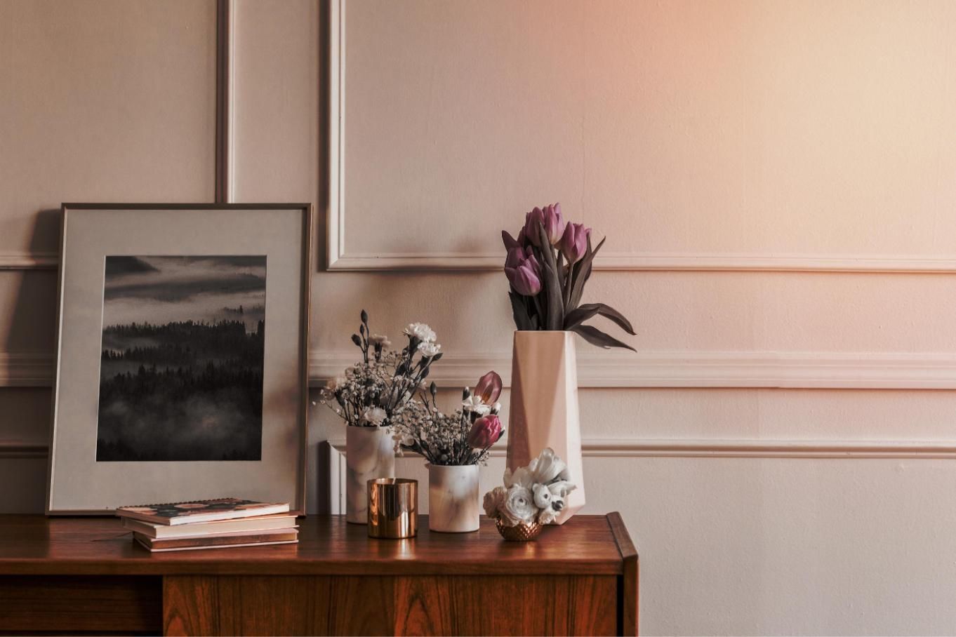 Armario de madera con flores, arte y libros contra una pared con moldura decorativa.